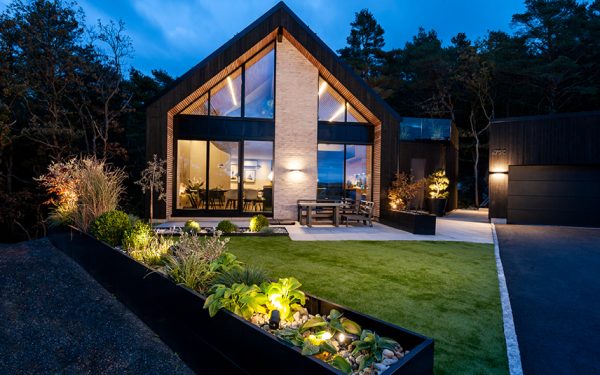 stor og moderne villa med store, heldekkende vindusflater og grønn hage med utebelysning i blomsterbed og på fasade.